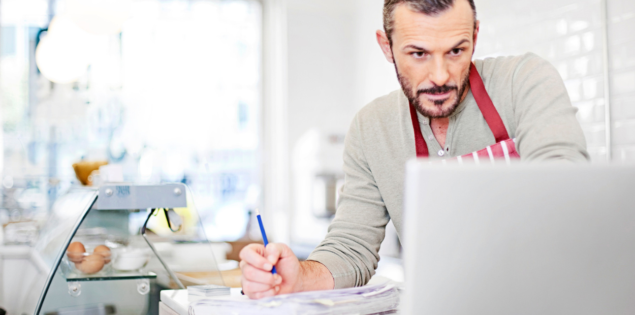 A white man with dark hair is working on a laptop. He is wearing an apron and working in a coffee shop.